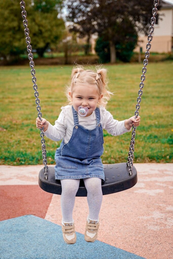Child with pacifier on swing, outdoors in a park.