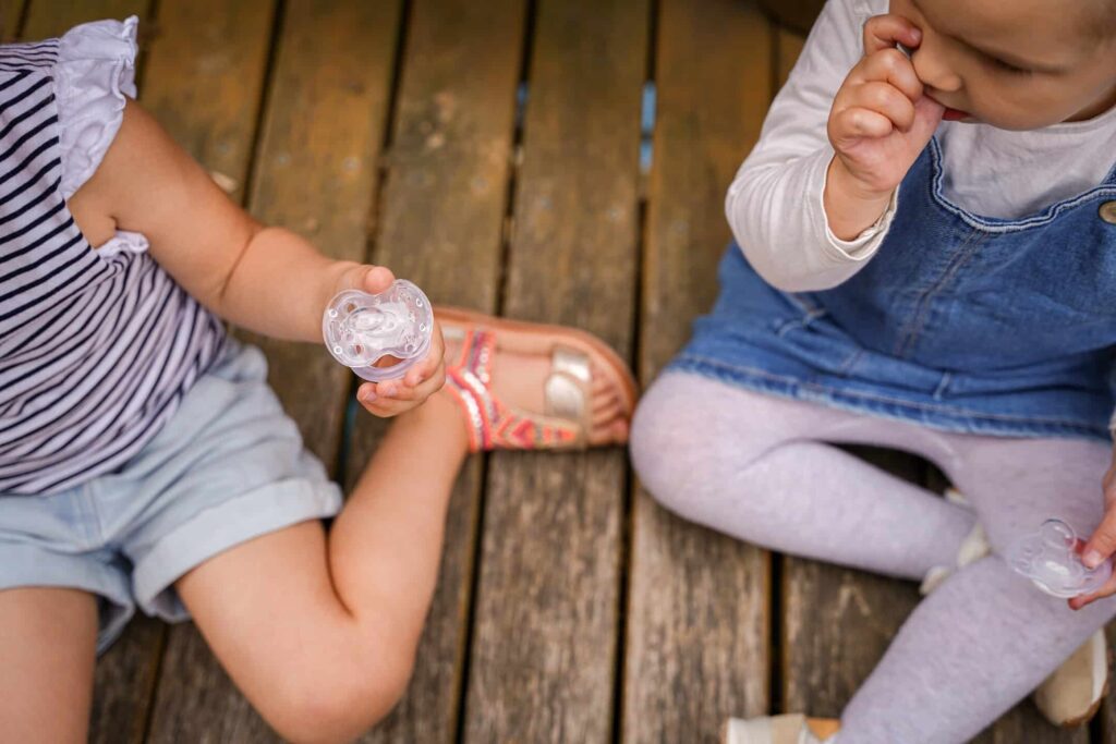 Two toddlers sitting with pacifiers on a wooden surface.